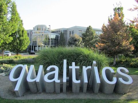 A set of three dimensional letters coming out the ground to spell Qualtrics amongst some brush in front of an office style building