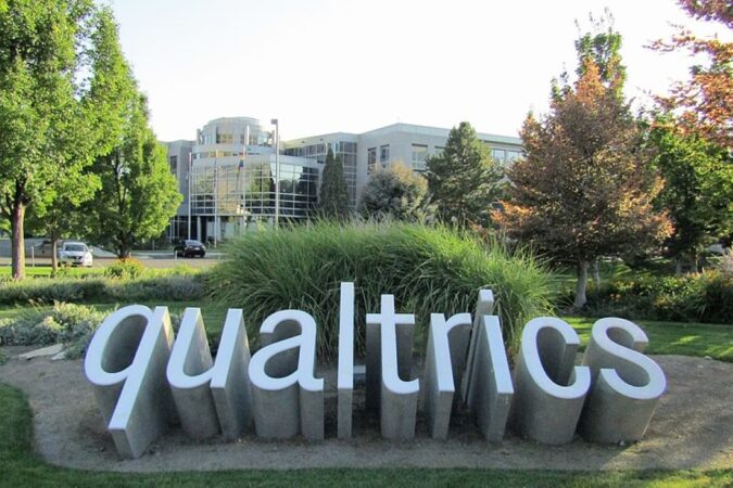 A set of three dimensional letters coming out the ground to spell Qualtrics amongst some brush in front of an office style building