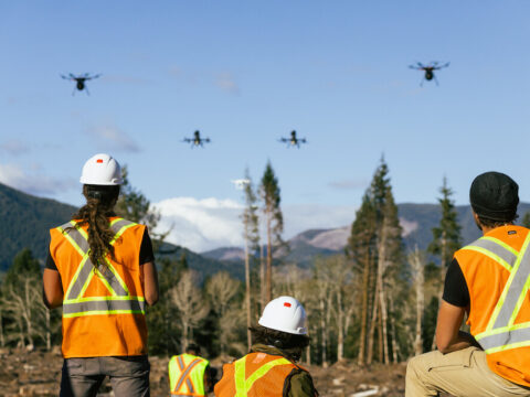 three workers in visibility vests observe as drones take off over burn landscape with mountains and forest in the background