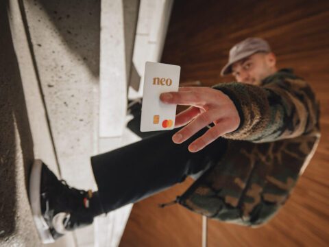 a man in skateboard shoes leans sideways and flashes a Neo mastercard at the camera it is in focus in the foreground and he is out of focus in the background