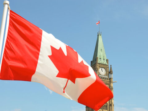 Canadian flag flies in the foreground over the parliament buildings in Ottawa on a sunny day