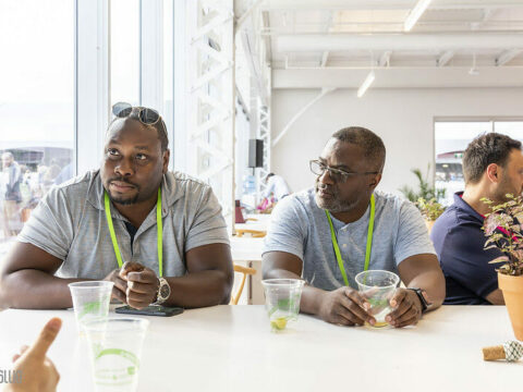 Two men wearing lanyards sit at a table with plastic cups in front of them at a past startupfest event