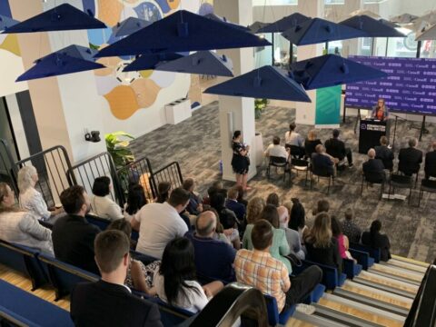 people sit in edmonton unlimited's event space watching a female speaker. cool lighting fixtures that look like umbrellas are overhead. there's a steeply tiered seating area and about 60-70 people in attendance