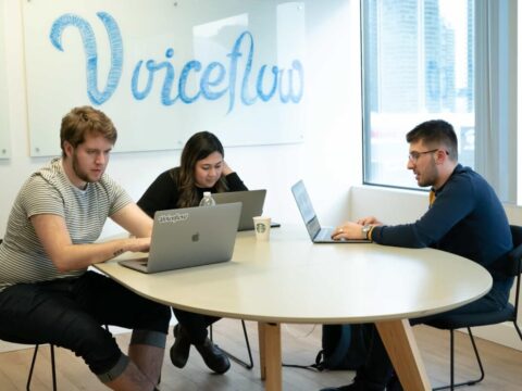 three people work at computers at a desk in a brightly lit office with the voiceflow logo on the wall