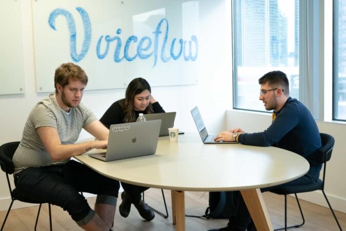 three people work at computers at a desk in a brightly lit office with the voiceflow logo on the wall