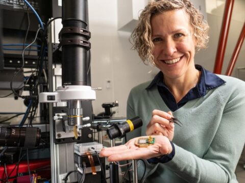 Photonic co-founder Stephanie Simmons holding a chip in her raised hand. She is smiling and appears to be in some kind of workshop.